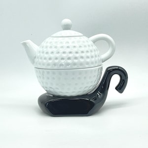 Op de afbeelding: Een witte keramische theepot met een golfbalontwerp, inclusief deksel en handvat. De theepot staat op een zwarte basis met een gebogen handvat. Ideaal voor golfliefhebbers.