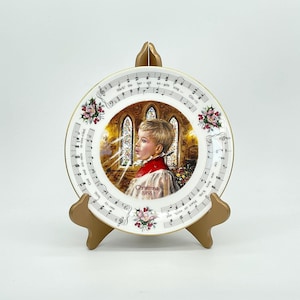 Könnte beinhalten: Ein weißer Keramik-Weihnachtsteller mit goldener Verzierung und einem Bild eines jungen Jungen, der in einer Kirche singt. Der Teller hat Noten am Rand und die Worte "Weihnachten 1988".