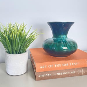 Puede incluir: Un jarrón de cerámica esmaltado en verde y azul con una boca ancha y un cuerpo redondeado. El jarrón está colocado sobre dos libros, uno naranja y otro marrón. Los libros tienen los títulos "Art of the Far East" y "African and Oceanic Art".