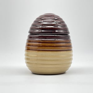 Może przedstawiać: Gliniany słoik na miód w kolorze brązowym i beżowym z pokrywką. Słoik ma kształt ula i ma fakturowaną powierzchnię.