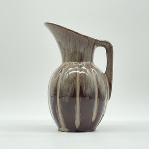 Puede incluir: Una jarra de cerámica marrón y crema con un diseño a rayas. La jarra tiene un cuerpo redondo y un asa curva.