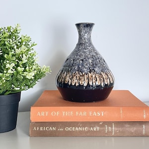 May include: A ceramic vase with a dark brown base, a textured grey and black middle section, and a narrow neck. The vase sits on two books with orange and brown covers. The books are titled "ART OF THE FAR EAST" and "AFRICAN AND OCEANIC ART."