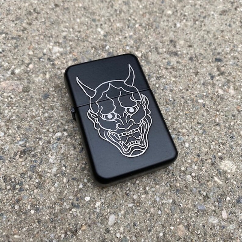 May include: Black lighter featuring a white outline of a Japanese demon mask.