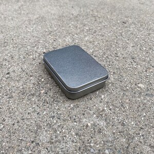 May include: A small, rectangular, silver tin box with a textured surface.