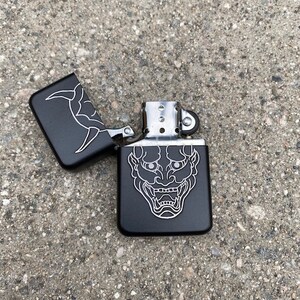May include: Black lighter with a silver hinge and a white outline of a Japanese demon face on the front.