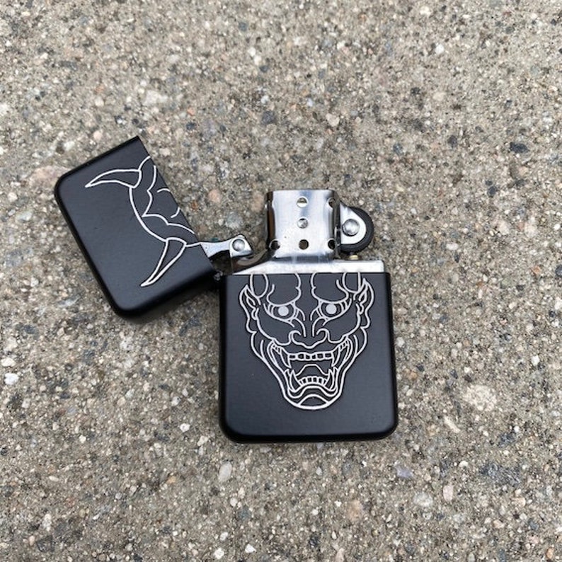 May include: Black lighter with a silver hinge and a white outline of a Japanese demon face on the front.