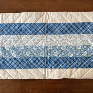 May include: A quilted table runner with a white background and blue and white patterned stripes. The runner has a diamond quilting pattern.