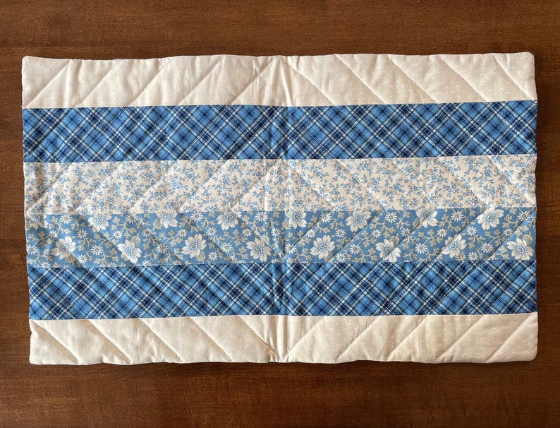 May include: A quilted table runner with a white background and blue and white patterned stripes. The runner has a diamond quilting pattern.
