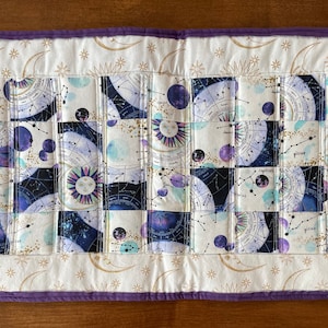 Könnte beinhalten: Gesteppter Tischläufer mit einem Himmelsdesign. Die Mitte hat ein Rastermuster mit Tierkreiszeichen, Planeten und Sternbildern in Blau-, Lila- und Weißtönen. Der Rand ist cremefarben mit goldenen Monden und Sternen und einem lila Rand.