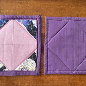 Puede incluir: Dos agarraderas acolchadas de color morado. La de la izquierda tiene un tejido rosa en forma de diamante en el centro con un estampado de constelaciones en los bordes exteriores. La de la derecha es de color morado sólido.