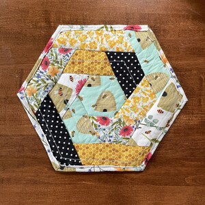 May include: Hexagon-shaped quilted coaster with a beehive and floral pattern. The coaster is made of fabric with a variety of colors, including yellow, black, white, green, and red.