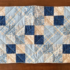 May include: A blue and cream patchwork quilt runner with a diamond pattern. The runner is made of various fabrics with floral and plaid designs.