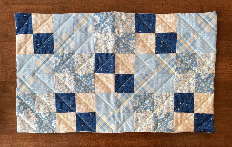 May include: A blue and cream patchwork quilt runner with a diamond pattern. The runner is made of various fabrics with floral and plaid designs.