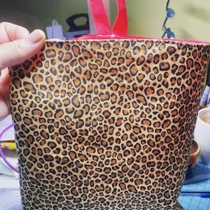 May include: A tote bag with a leopard print pattern in shades of brown and black. The bag has a red interior and red ribbon handles. The bag is held by a hand.