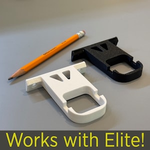 May include: Two 3D printed wall mounts, one white and one black, designed to hold a curtain rod. The mounts are shown on a gray surface with a yellow pencil. The text "Works with Elite!" is visible at the bottom of the image.