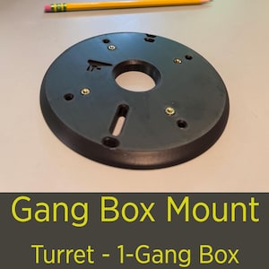 May include: A black, circular Gang Box Mount with a central hole and several smaller holes for screws. The mount has a flat, matte finish and includes the text "Gang Box Mount" and "Turret - 1-Gang Box" in yellow. A pencil rests above the mount.