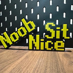 Pode incluir: Dois sinais pixelados amarelos e pretos com as palavras "Noob" e "Sit Nice" em uma superfície de madeira marrom.