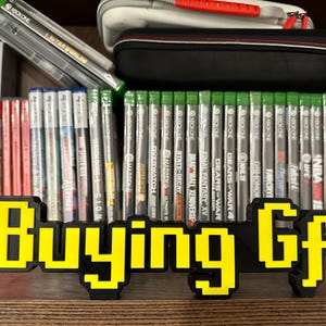 May include: A yellow and black pixelated sign that reads "Buying Gf" in front of a shelf of video games. The games are all for the Xbox One console.