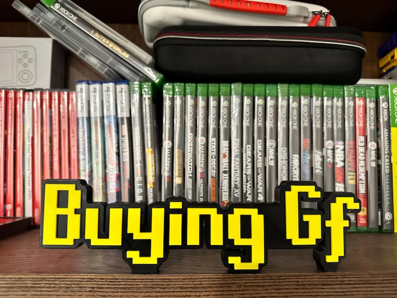 May include: A yellow and black pixelated sign that reads "Buying Gf" in front of a shelf of video games. The games are all for the Xbox One console.