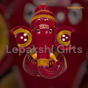 May include: A red wooden figurine of the Hindu deity Ganesha, with yellow and white painted details. The figurine is decorated with a red and yellow striped headdress and has a small, white painted third eye on its forehead.