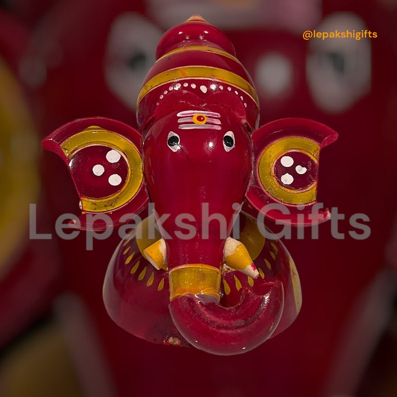 May include: A red wooden figurine of the Hindu deity Ganesha, with yellow and white painted details. The figurine is decorated with a red and yellow striped headdress and has a small, white painted third eye on its forehead.