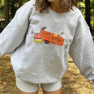 May include: A light grey crewneck sweatshirt featuring an embroidered Halloween design. The graphic includes a candy corn, a small spider, a yellow star, a white ghost, and an orange rectangular patch with the text trick or treat.