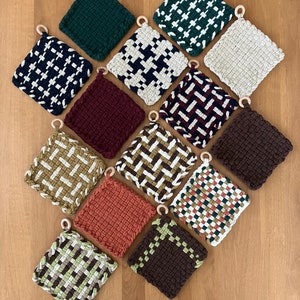May include: A collection of 12 woven pot holders in various colors and patterns, including green, white, black, red, brown, tan, and orange. Each pot holder has a wooden ring attached for hanging.