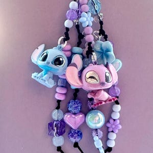 May include: A keychain featuring two cartoon character charms, one blue and one pink, with beaded accents in shades of purple, blue, and white. The charms are attached to a silver-colored clasp.