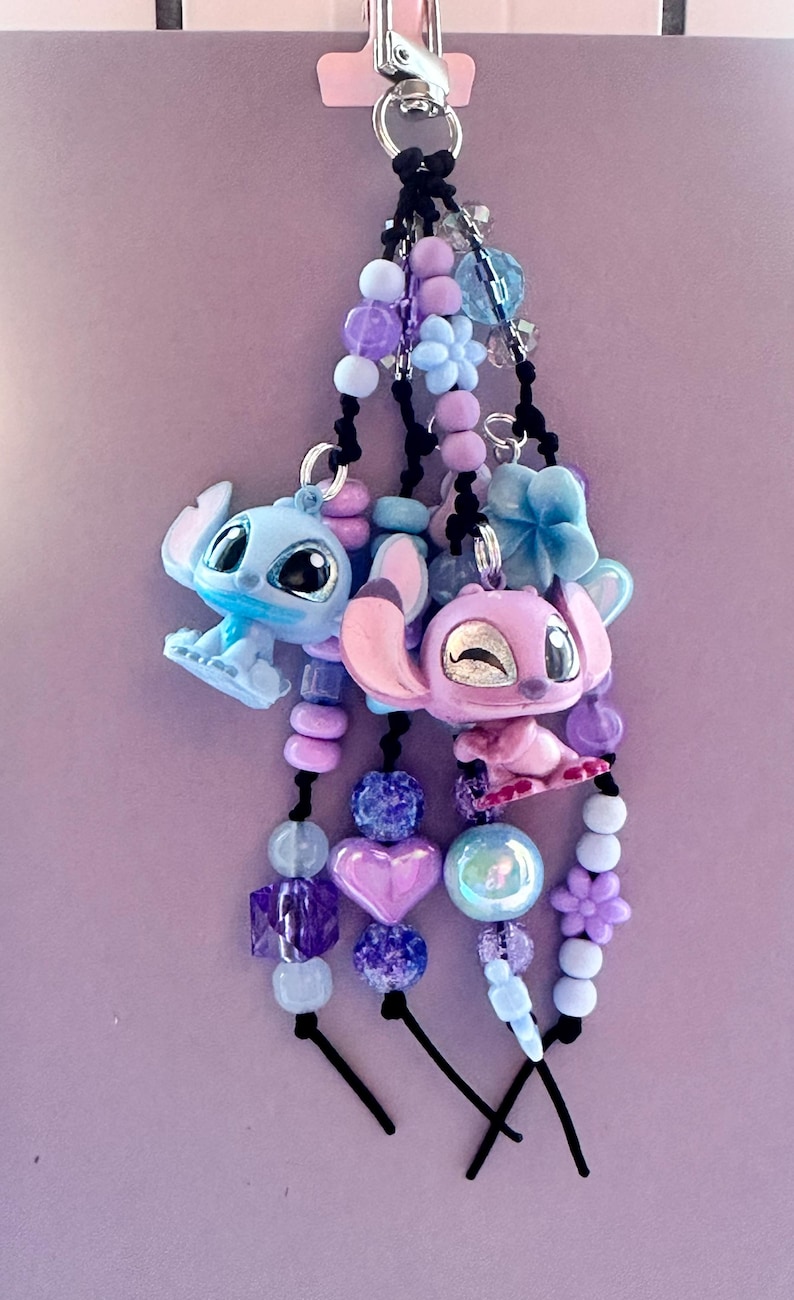 May include: A keychain featuring two cartoon character charms, one blue and one pink, with beaded accents in shades of purple, blue, and white. The charms are attached to a silver-colored clasp.
