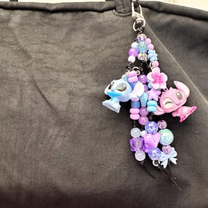 May include: A beaded charm featuring a blue and pink cartoon character, with a variety of purple, blue, and pink beads. The charm is attached to a black bag with a silver clasp. The charm includes flower and star accents.