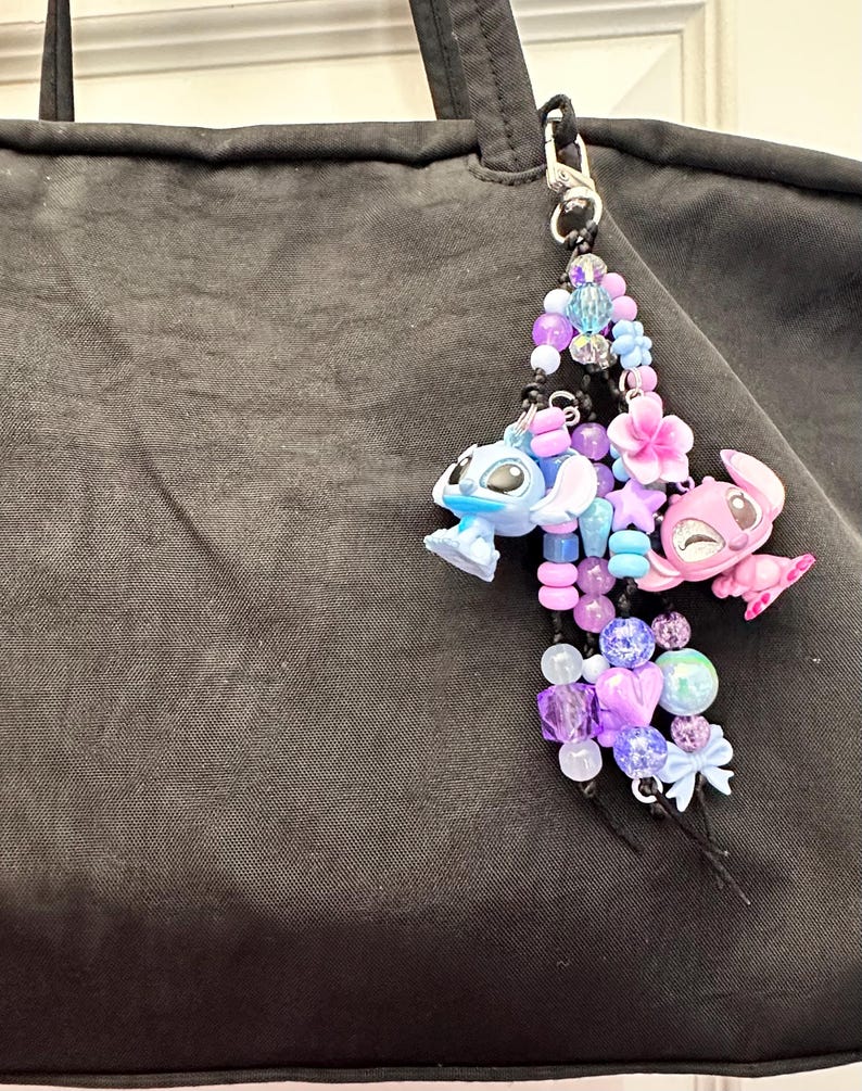 May include: A beaded charm featuring a blue and pink cartoon character, with a variety of purple, blue, and pink beads. The charm is attached to a black bag with a silver clasp. The charm includes flower and star accents.