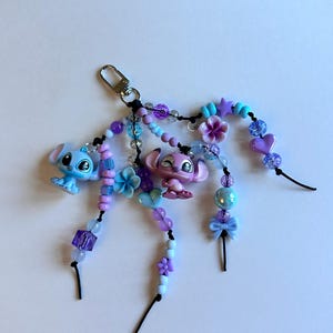 May include: A handmade keychain featuring two cartoon character figurines, one blue and one pink, with beaded accents in shades of purple, blue, and white. The keychain includes a silver-colored clasp and decorative beads, flowers, and bows.