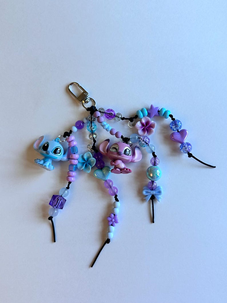 May include: A handmade keychain featuring two cartoon character figurines, one blue and one pink, with beaded accents in shades of purple, blue, and white. The keychain includes a silver-colored clasp and decorative beads, flowers, and bows.