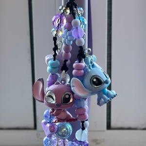 May include: A beaded keychain featuring two character figurines, one pink and one blue, with a variety of purple, blue, and clear beads. The keychain has a silver clasp and black string accents.