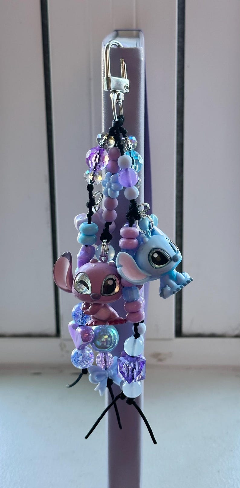 May include: A beaded keychain featuring two character figurines, one pink and one blue, with a variety of purple, blue, and clear beads. The keychain has a silver clasp and black string accents.
