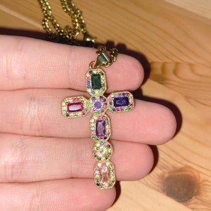 May include: A gold chain necklace with a multi-colored gemstone cross pendant. The cross features emerald, amethyst, ruby, and sapphire gemstones.