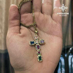 May include: A gold chain necklace with a multi-colored gem-encrusted cross pendant. The cross features rectangular-shaped gems in shades of pink, purple, blue, green, and red.