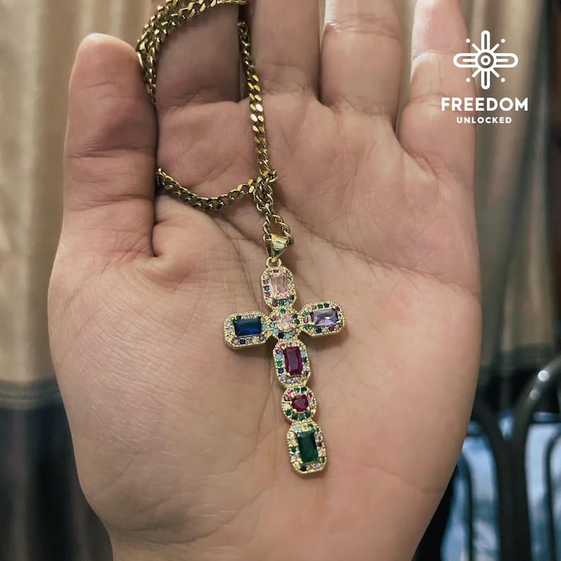 May include: A gold chain necklace with a multi-colored gem-encrusted cross pendant. The cross features rectangular-shaped gems in shades of pink, purple, blue, green, and red.