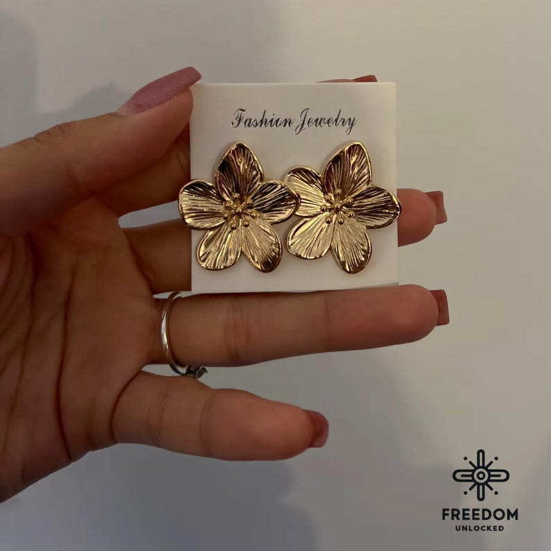 Op de afbeelding: Een paar gouden oorringen in de vorm van een bloem op een witte kaart met de tekst "Fashion Jewelry".