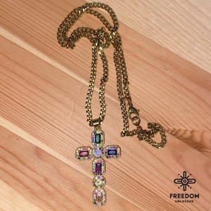May include: A gold chain necklace with a large, multi-colored jeweled cross pendant. The cross is adorned with various colored gemstones, including purple, pink, green, blue, and yellow.