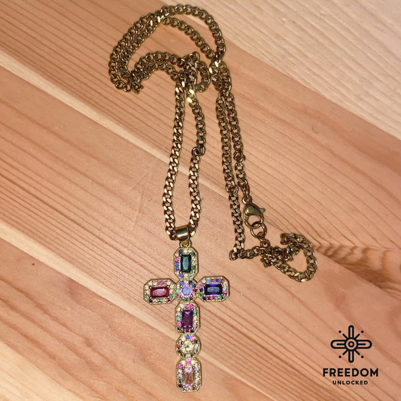 May include: A gold chain necklace with a large, multi-colored jeweled cross pendant. The cross is adorned with various colored gemstones, including purple, pink, green, blue, and yellow.