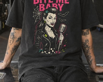 May include: A charcoal grey t-shirt featuring a graphic print of a vampire with fangs holding a vintage microphone. The text BITE ME BABY appears in pink above the character, accompanied by illustrations of a spider web and two small bats.