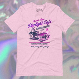 May include: Pink t-shirt with a retro-style graphic design for a cafe called "Cosmic Rays Starlight Cafe" in Tomorrowland. The design features a futuristic building with palm trees and the text "Burgers, Chicken & Fries. Best quality in the galaxy!"