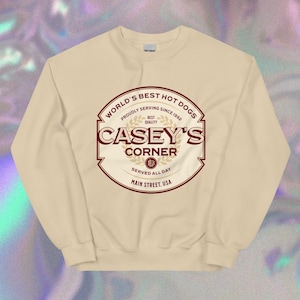 May include: A beige long-sleeved sweatshirt with a red and gold logo that says "World's Best Hot Dogs" and "Casey's Corner". The logo also includes the text "Proudly Serving Since 1992", "Best Quality", "Served All Day", and "Main Street, USA".