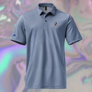 May include: A light blue polo shirt with a collared neckline and short sleeves. The shirt features a small Mickey Mouse embroidered graphic on the chest and the Adidas logo on the sleeve. The shirt has a three-button placket.