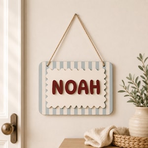 Op de afbeelding: Een rechthoekig wandbord met een lichtblauw en wit verticaal gestreepte achtergrond en een crèmekleurig middenpaneel met geschulpte randen. De naam NOAH staat in dikke, donkerrode, afgeronde hoofdletters. Een touw is bevestigd om op te hangen.