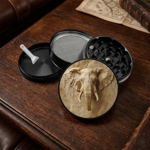 May include: A black multi-part herb grinder featuring a circular lid with a tan, paper-like elephant head graphic. The set includes a metal mesh screen, a black base compartment, and a small white plastic scraper tool for processing dry herbs.