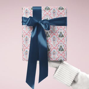May include: A wrapped gift with a navy blue satin ribbon and bow. The wrapping paper has a repeating pattern of nutcrackers, Christmas trees, and pink bows on a white background. The gift is held by a person wearing a white glove.