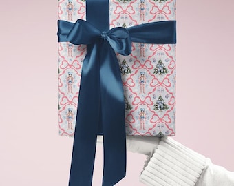 Peut inclure: Un cadeau emballé avec un ruban et un nœud en satin bleu marine. Le papier d'emballage présente un motif répété de casse-noisettes, de sapins de Noël et de nœuds roses sur fond blanc. Le cadeau est tenu par une personne portant un gant blanc.