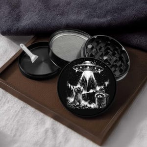 May include: Black and white herb grinder with a raccoon and UFO design. The grinder is disassembled, showing the different parts, including a sifter and a small scoop. The design features a UFO beaming light onto two raccoons.